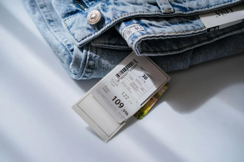 Label harga dan ukuran asli menempel pada celana jeans biru di atas meja putih, menunjukkan kondisi barang BNWT brand new with tags